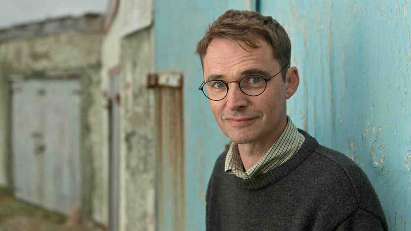 Dylan stands in front of boat sheds looking into the camera.