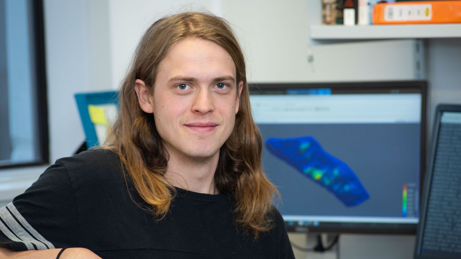 Clarrie sits at a desk looking directly at the camera. Behind him, his computer screen shows models of a glacier. Image copyright Victoria University of Wellington.