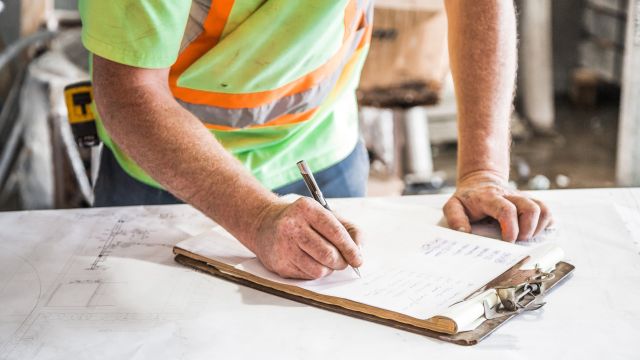 A tradesperson on a building site writes on a clipboard.