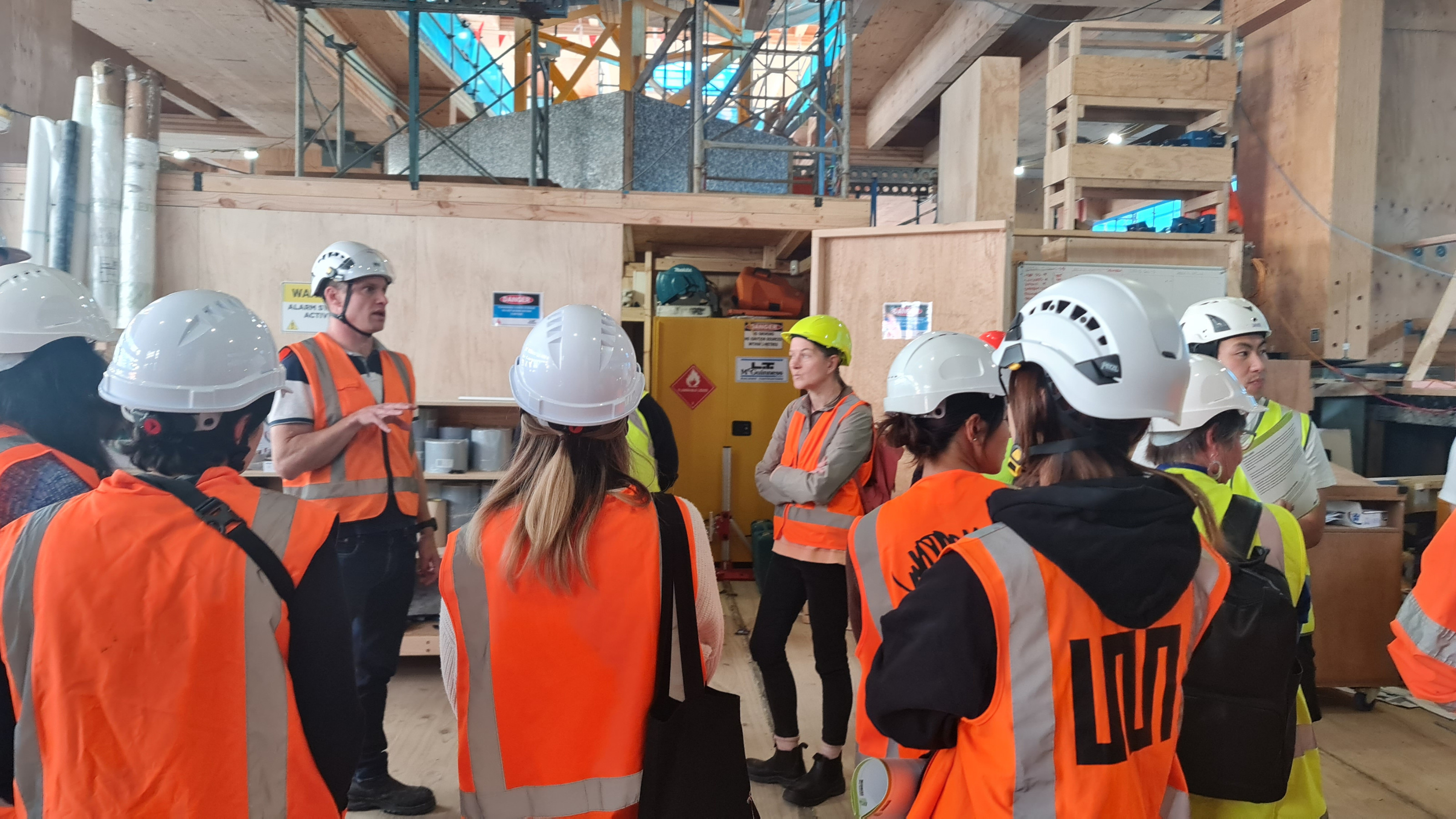 A group of people in hi vis vests and hard hats standing  listening to someone talk on a construction site.