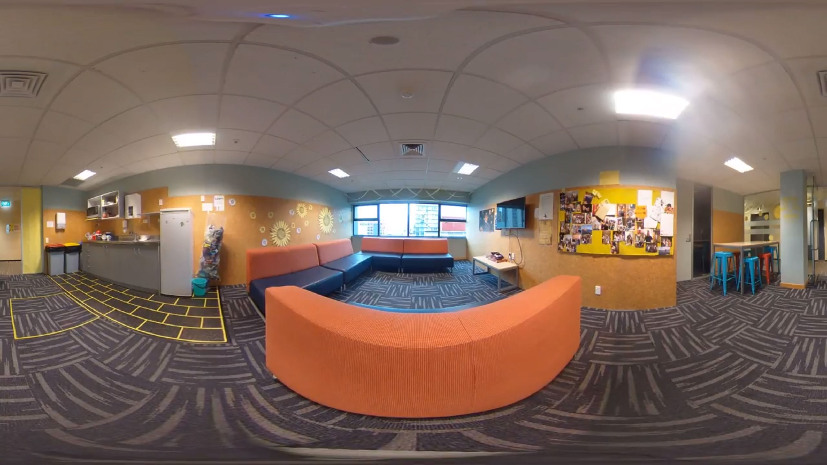 A panoramic view of a common space with orange sofas and a television.