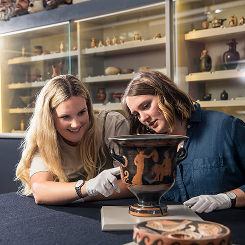 Two white gloved women interpret the red figure painting on a presumably ancient vase of Greek origin.