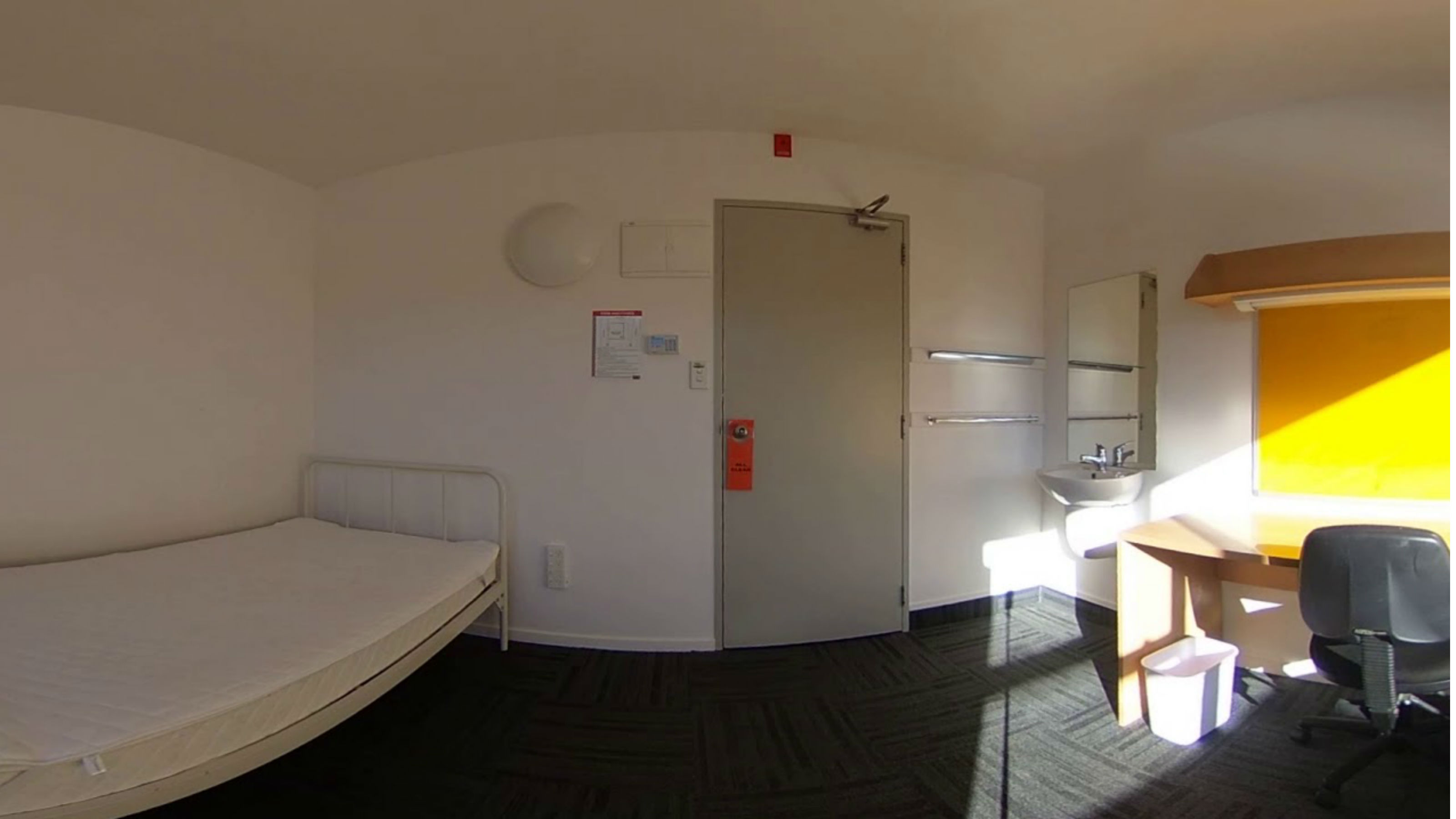 An empty bedroom with a single bed, a sink and mirror, and a desk with a chair.