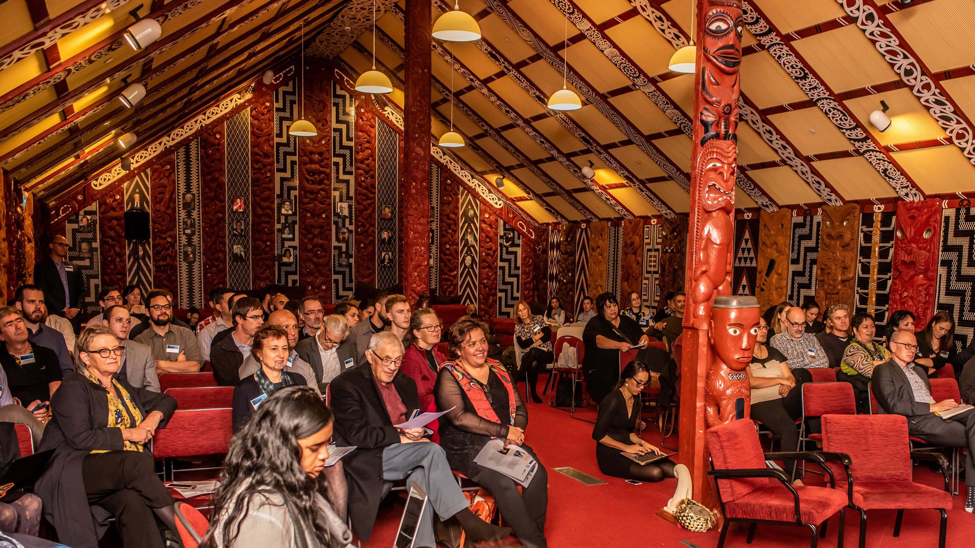 A large gathering of people sheltered within the carved wooden skeleton of the wharenui of Te Herenga Waka Marae.