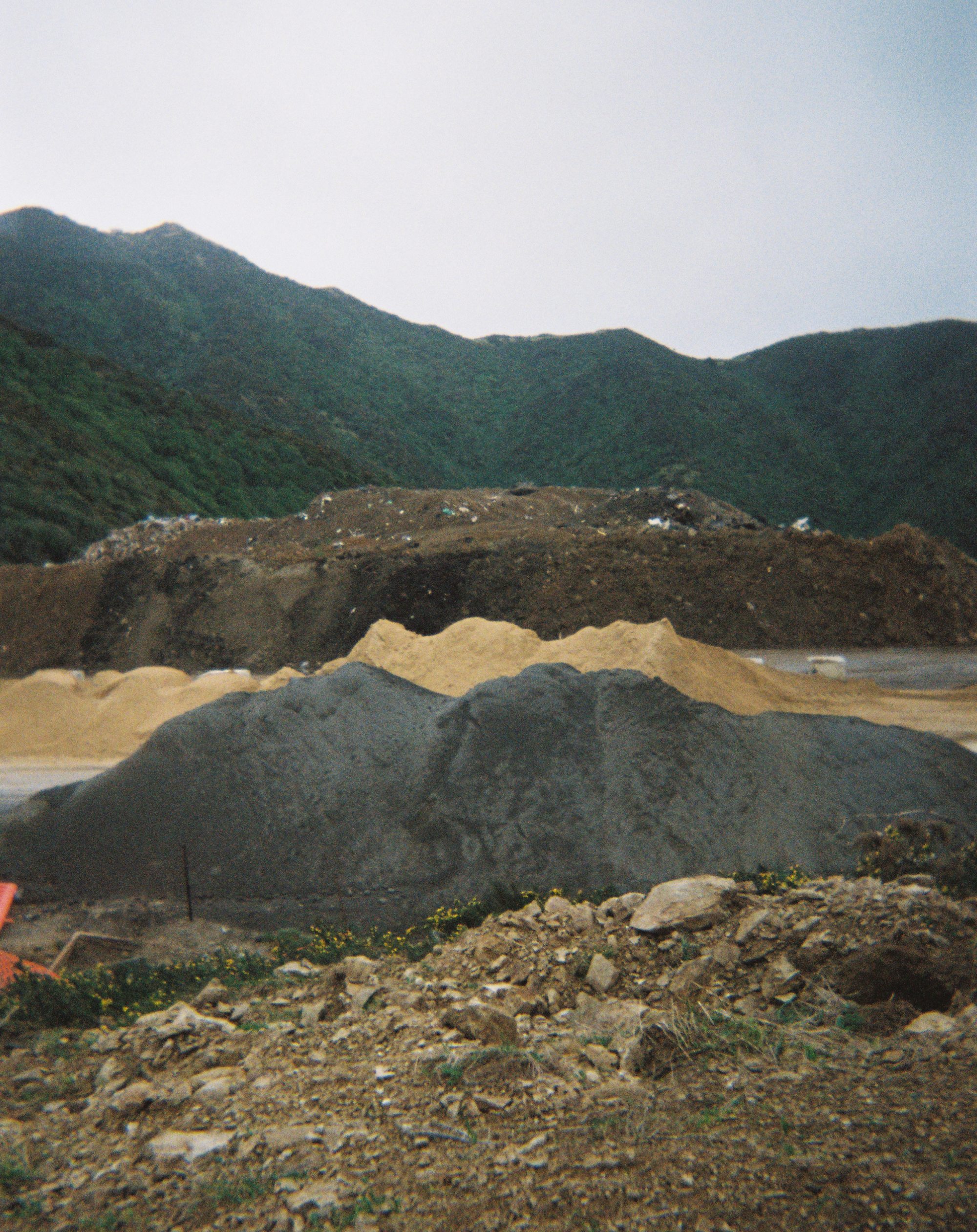 Green hills in the background with a foreground of piles of rubbish and various tones of gravel.