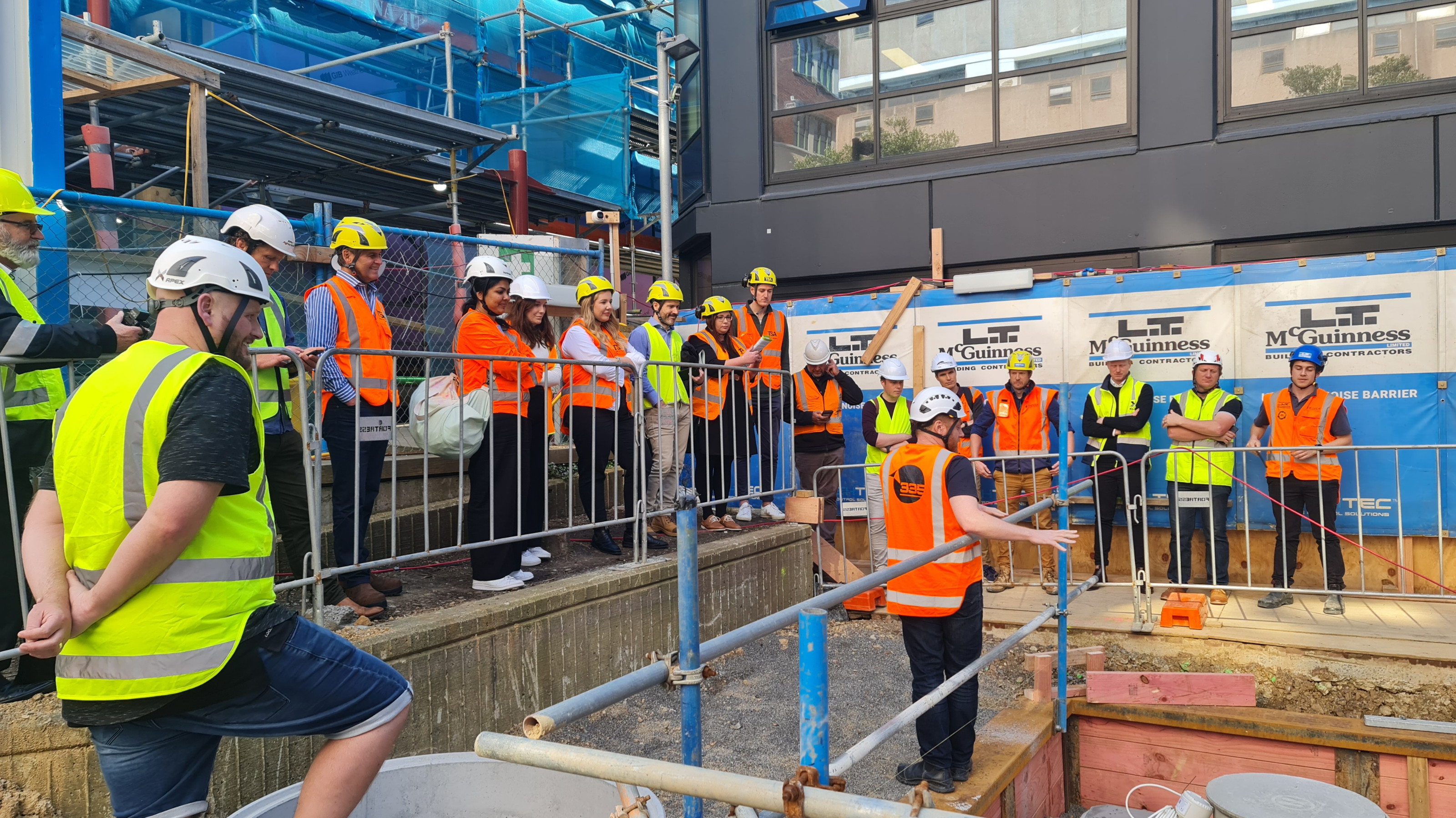 A group of people in hi vis vests and hard hats standing listening to someone talk at a wastewater treatment construction site.