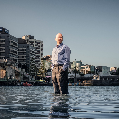 Dr Ilan Noy Man standing knee-deep in water, with city landscape in the background.