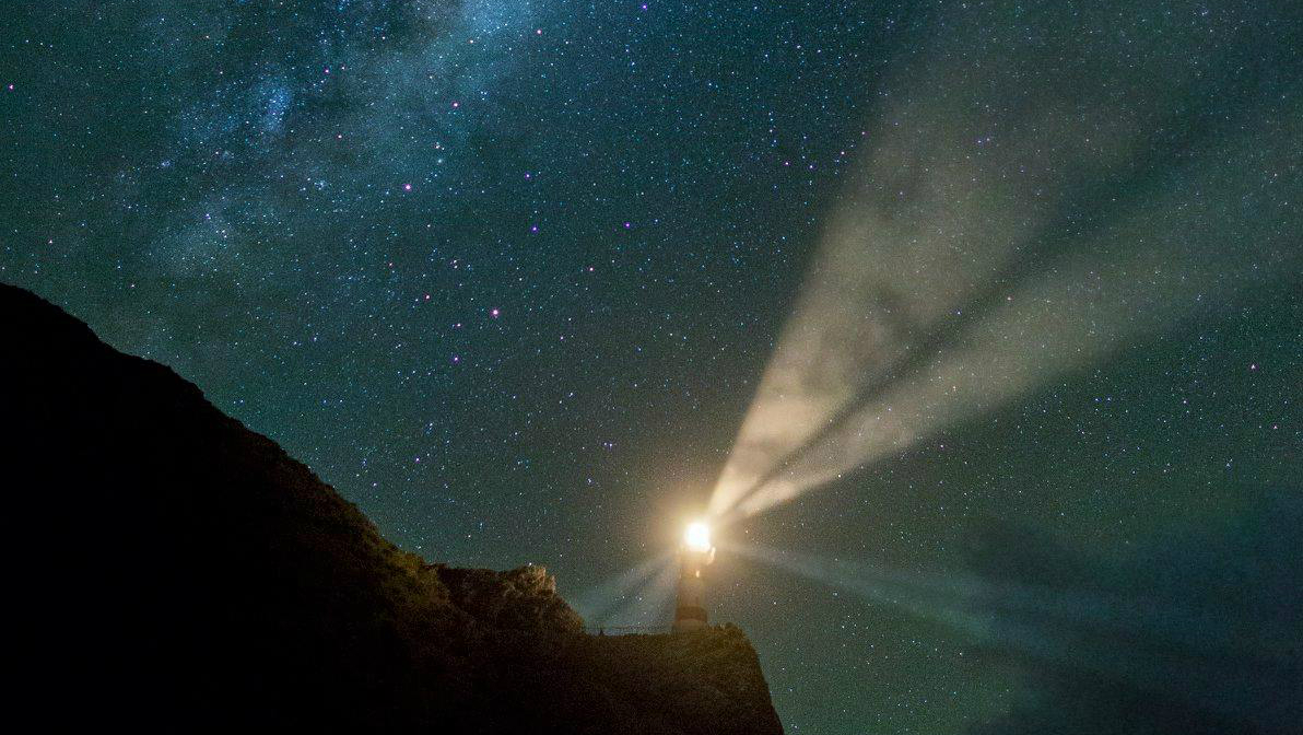 A lighthouse at night with a beam of light and the milky way above.