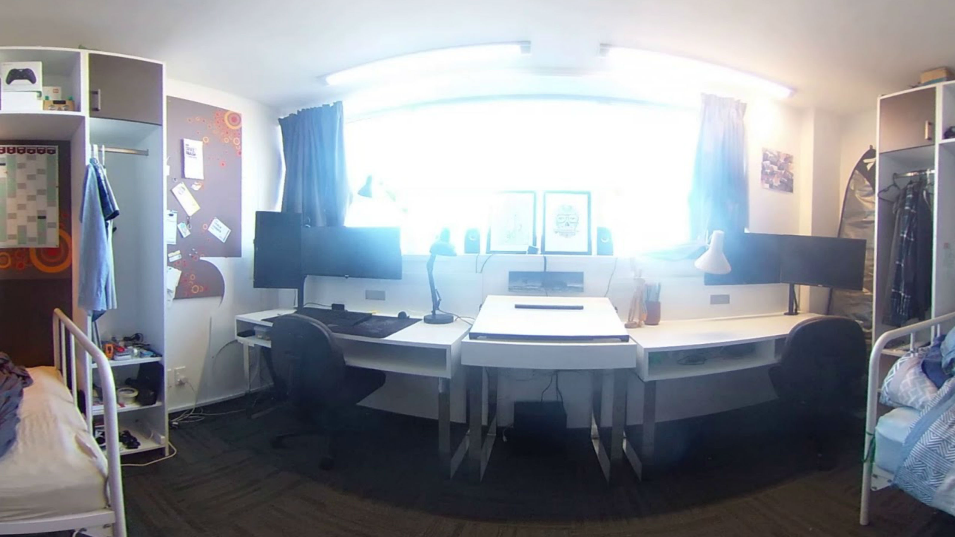 Image of a shared room with two desks that have multiple monitors, two wardrobes, and two beds.