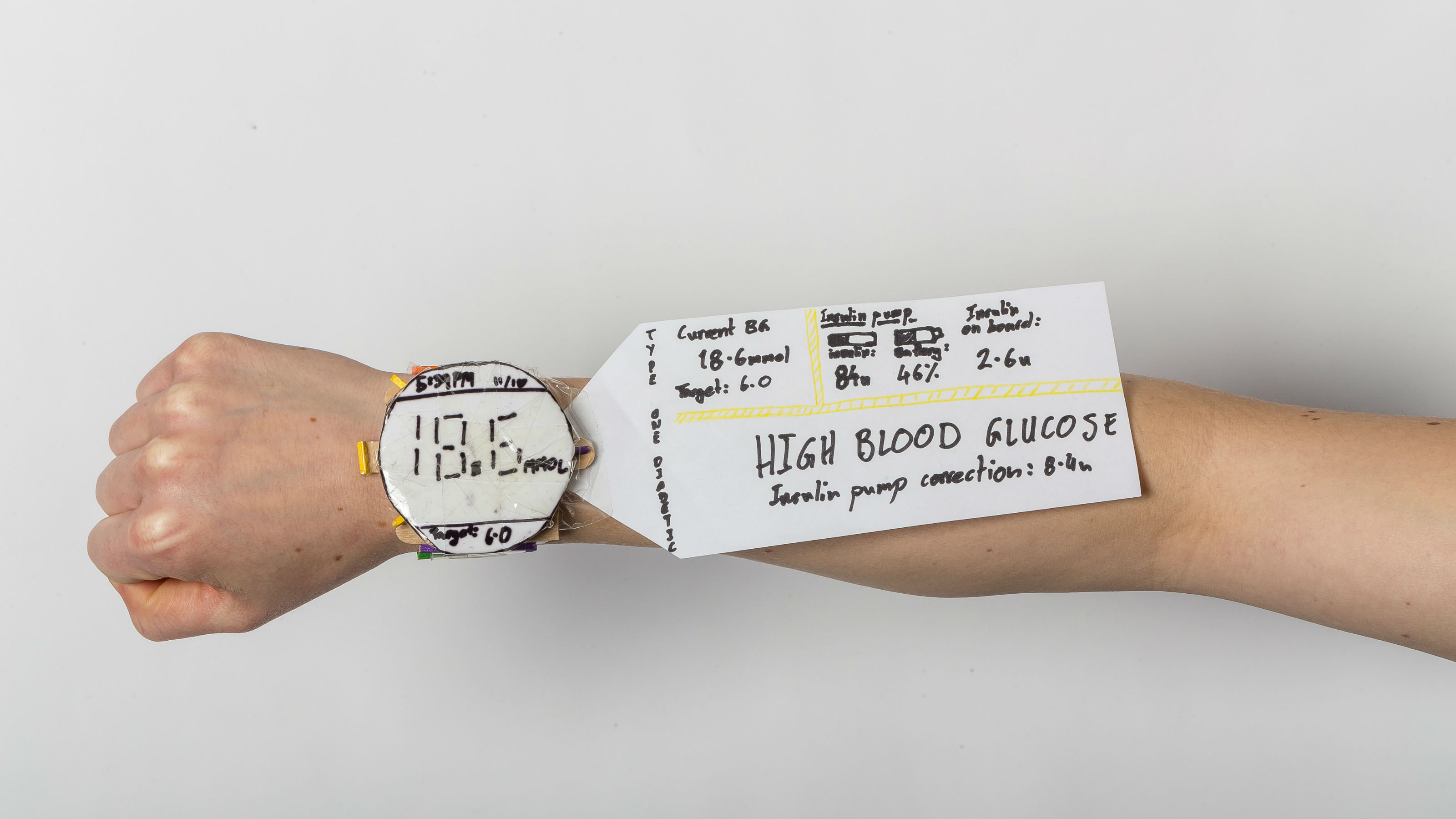 Arm wearing a watch design constructed of paper, with a paper information panel emerging from the side. Rapid paper prototype of a holographic watch concept created by design workshop participants (designers and adolescents with type 1 diabetes). Photo by Saint Andrew Matautia.
