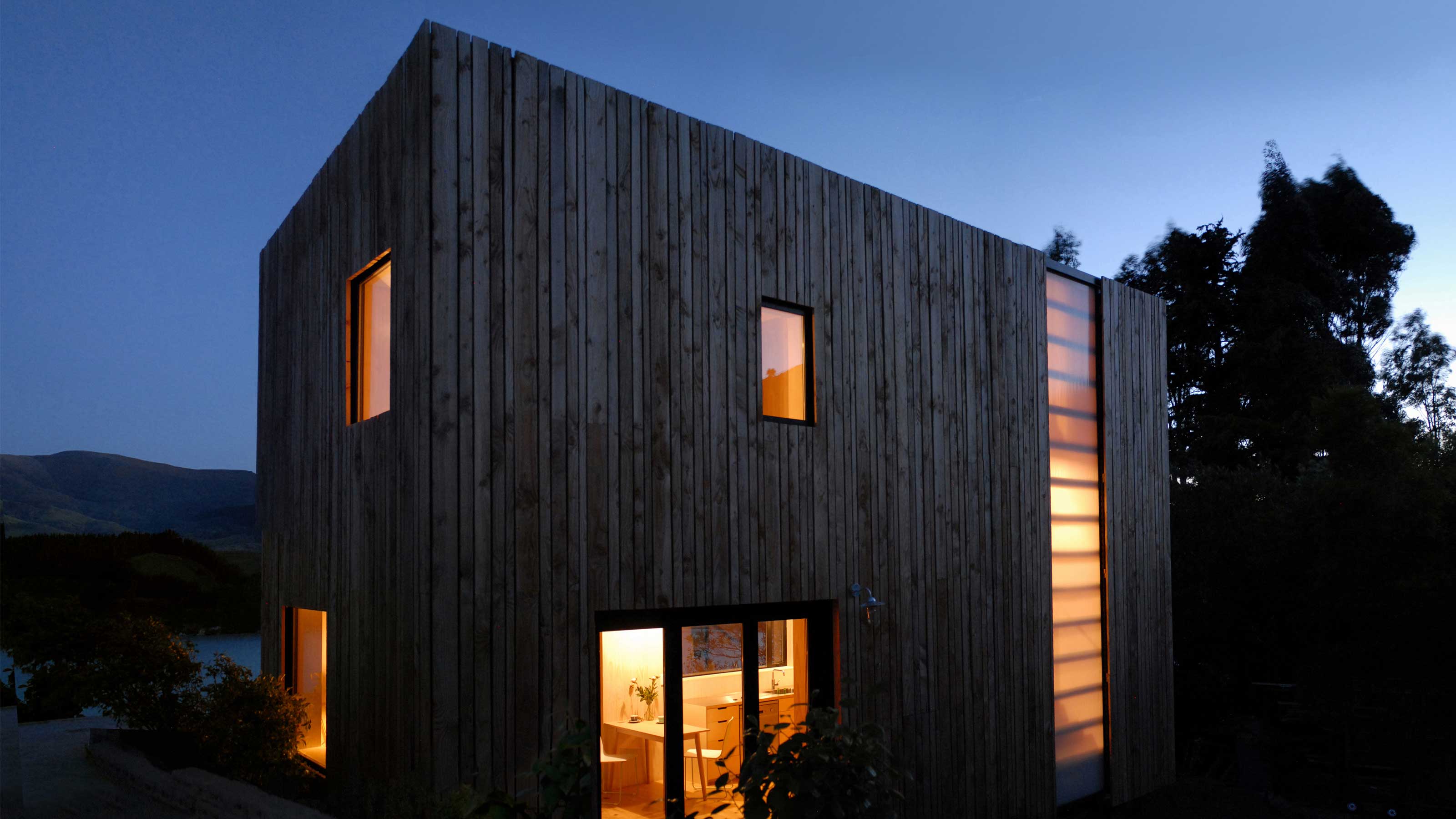 An image of the outside of a small residential building with orange lit windows at dusk.