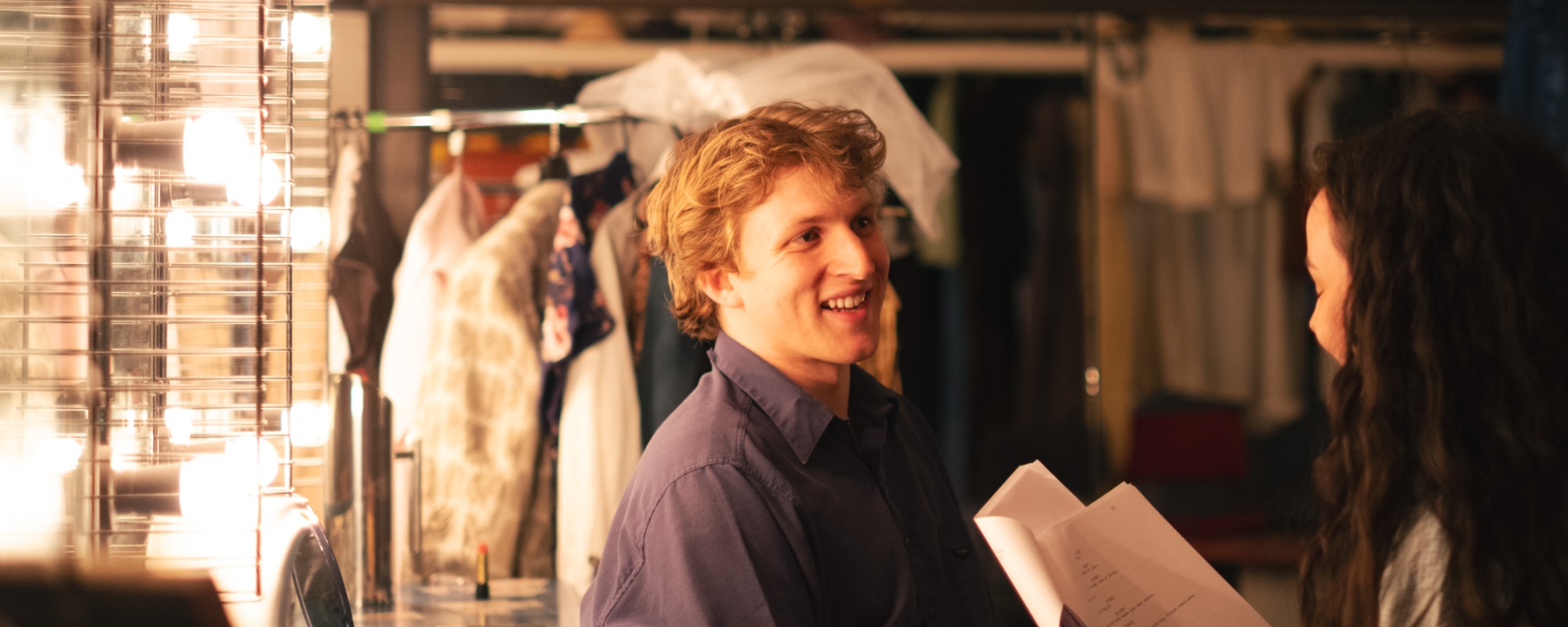 Two theatre students backstage in a greenroom, one of them holding a script.