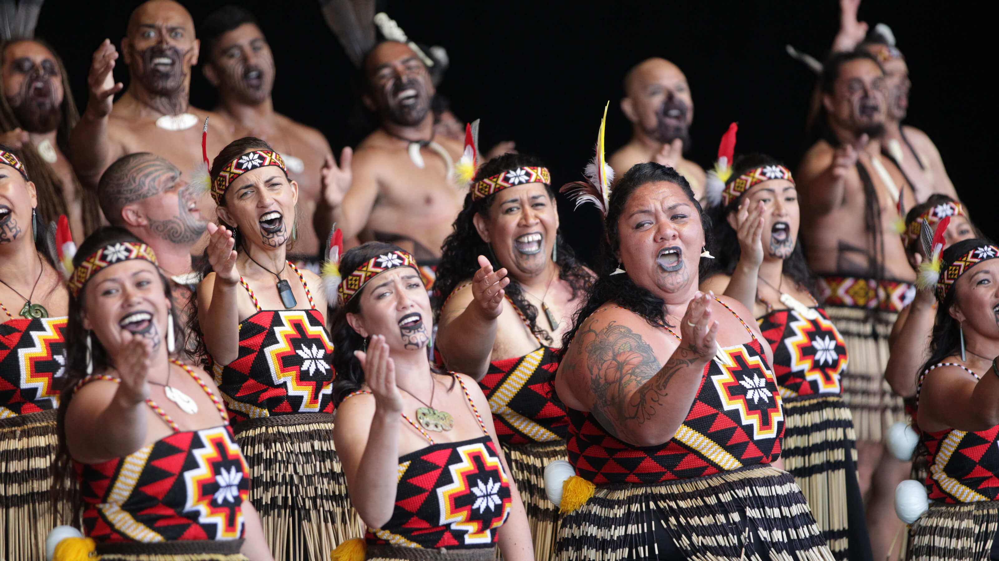 A Polynesian kapa haka group performing in tight, tiered lines and powerfully adorned with woven garments and carved pendants. Their figures twist and hands gesture in unison. They’re singing in chorus, and some deep-felt, collective emotion wells across their faces. 