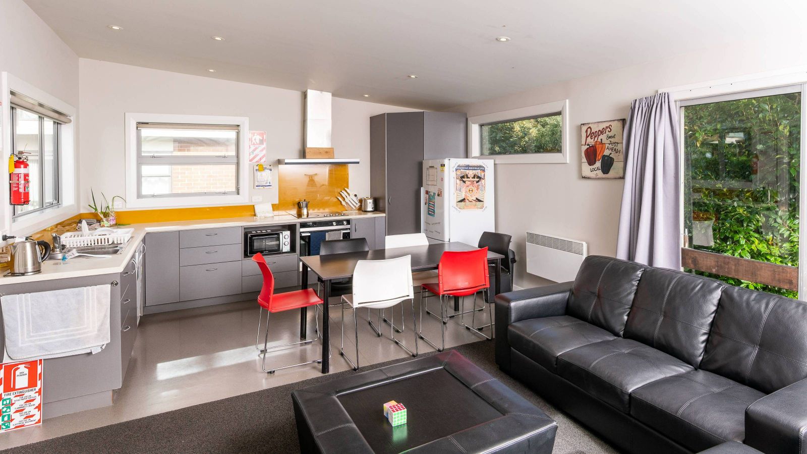 An open plan kitchen/living room with black leather couches and coffee table, grey kitchen counters, and a black dining table with red, white, and black chairs.