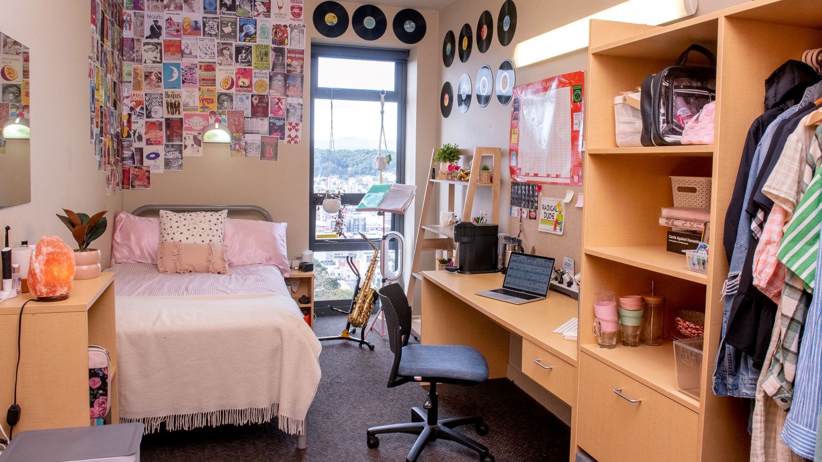 A modern, single room at Te Puni Village, with a bed next to a tall window, a desk, chair and wardrobe unit, and lots of pictures on the walls.