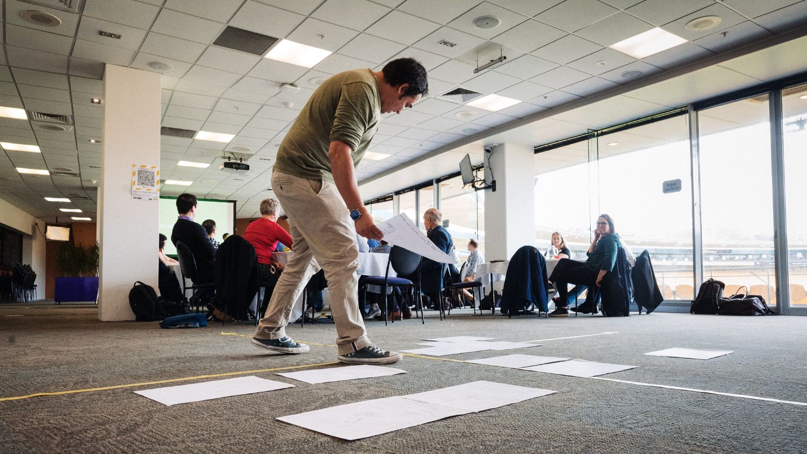 The person in the foreground is placing a piece of paper on the floor among other papers as people sit around round tables in the background