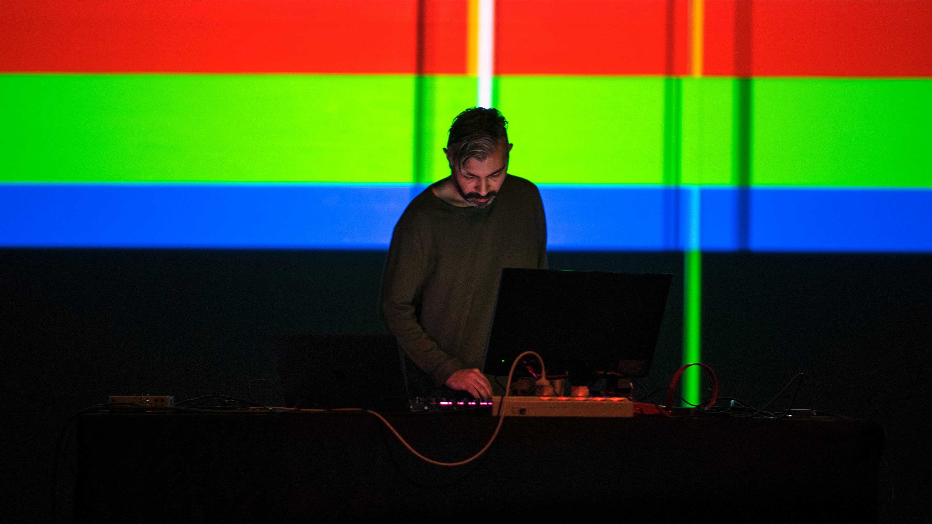Dr Mo Zareei performing a DJ set, with a blue, red, and green striped background. The lighting is low and moody.