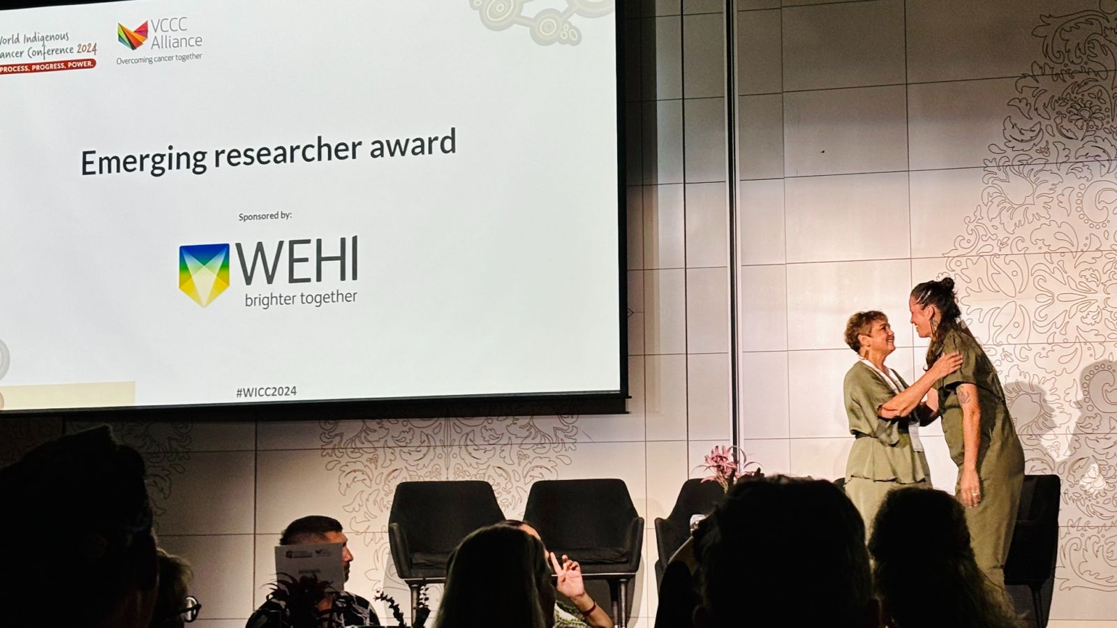 One person warmly greeting another on stage with a screen visible to the left announcing an award.