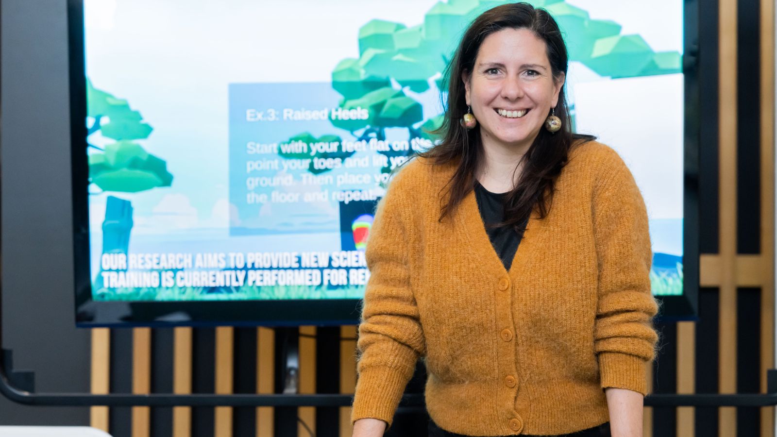 A woman is standing at a desk smiling at the camera. Behind her is a digital screen displaying gait training technology.