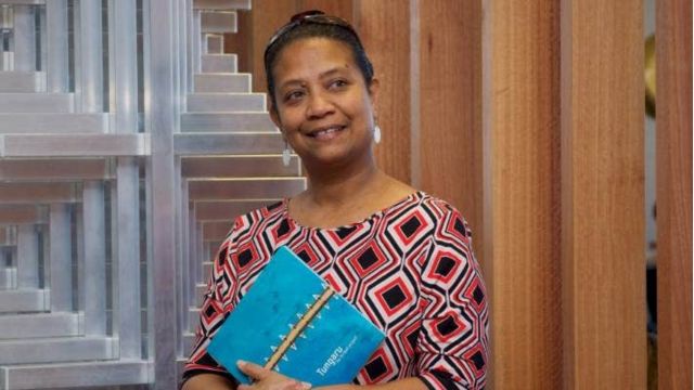 woman with dark hair pulled back, wearing colourful top, against background of wood and steel