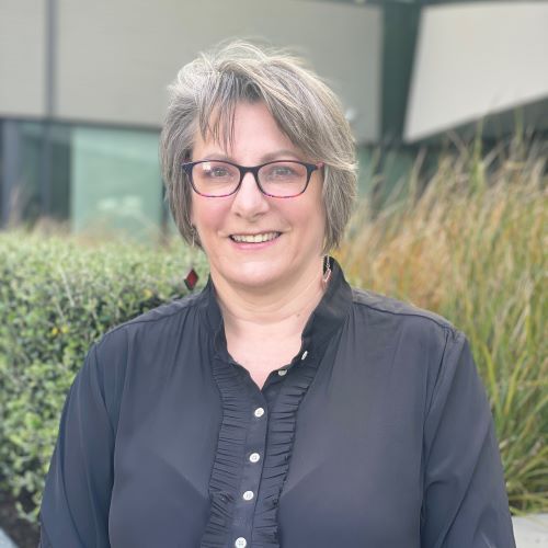 Woman wearing glasses standing and smiling at the camera. There is vegetation behind her