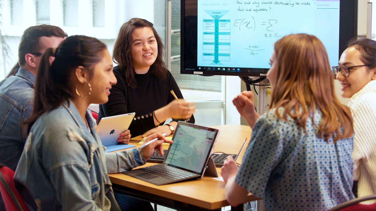 A group of students working together with large computer screen in the background.